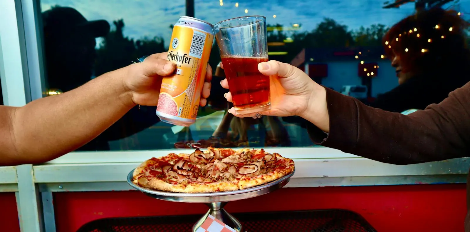 Two friends toast with a beer and a canned drink over a pizza on a stand