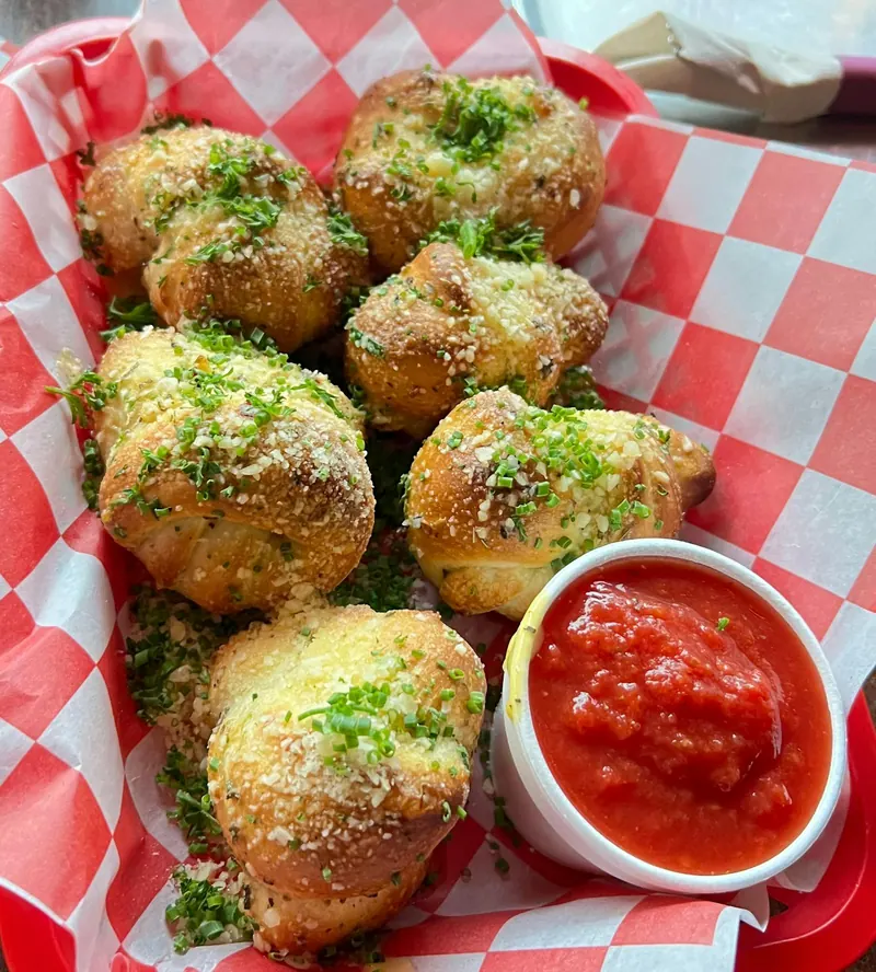 A basket of garlic knots topped with parmesan and chives, with a cup of marinara